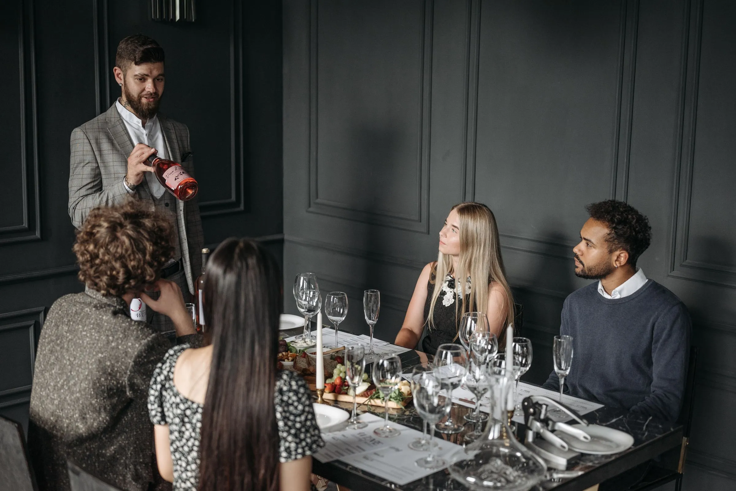 A wine host presenting a bottle to guests at a table